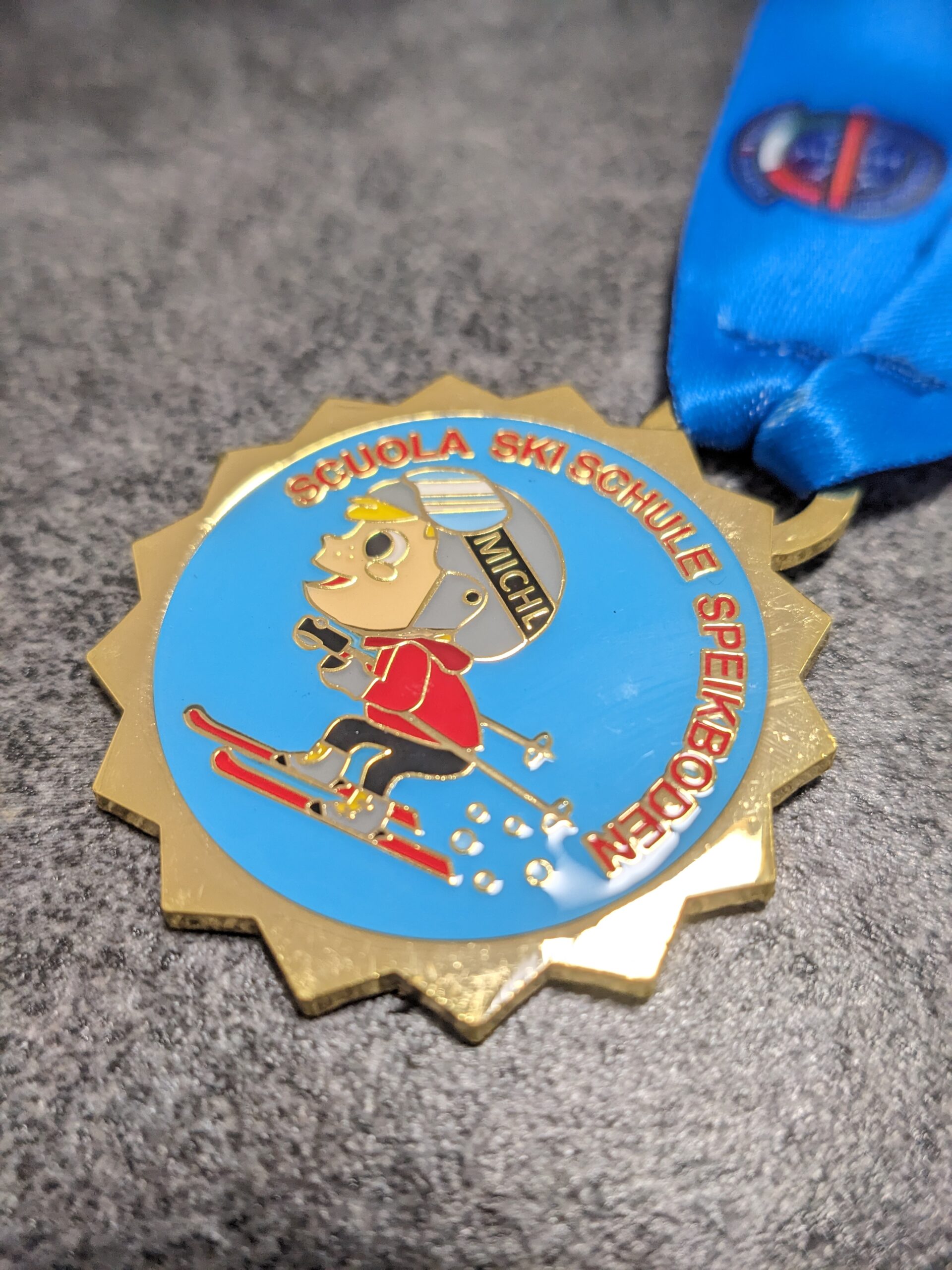 custom skischool speikboden medal in 2D gold finish enameled metal with lens effect and personalized sublimation ribbon side view custom skischool speikboden medal in 2D gold finish enameled metal with lens effect and personalized sublimation ribbon side view